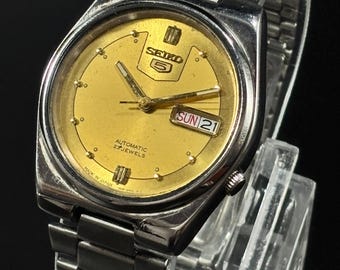 May include: A vintage Seiko 5 automatic wristwatch with a gold-colored face and a stainless steel band. The watch face displays the day and date, with the day reading "SUN 21". The watch is marked "Made in Japan" and "23 Jewels".