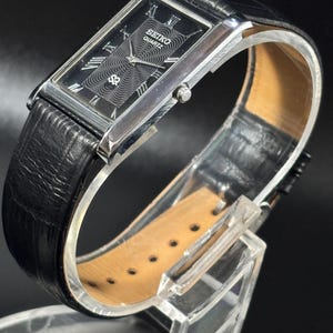 Puede incluir: Un reloj Seiko Quartz rectangular con esfera negra, caja plateada y correa de cuero negro. La esfera del reloj presenta n&uacute;meros romanos y un dise&ntilde;o estampado. El reloj se exhibe sobre un soporte transparente.
