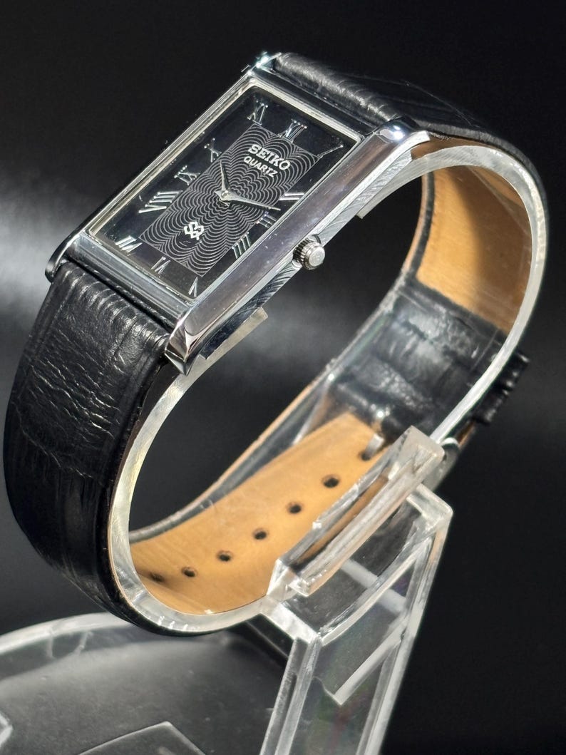 Puede incluir: Un reloj Seiko Quartz rectangular con esfera negra, caja plateada y correa de cuero negro. La esfera del reloj presenta n&uacute;meros romanos y un dise&ntilde;o estampado. El reloj se exhibe sobre un soporte transparente.