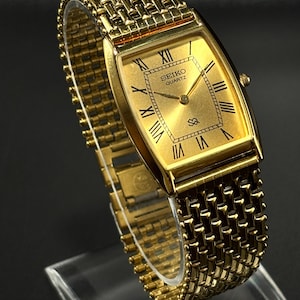 May include: A gold-toned Seiko Quartz watch with a rectangular face and Roman numeral hour markers. The watch features a mesh-style band and a gold dial. The watch is displayed on a clear stand.