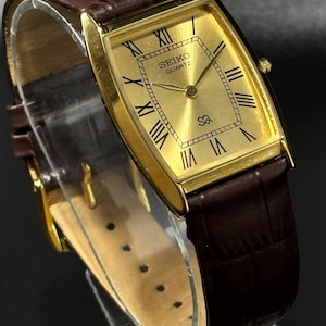 Może przedstawiać: Złoty zegarek Seiko Quartz z prostokątną tarczą i rzymskimi cyframi. Pasek jest brązowy, skórzany, ze wzorem krokodyla. Tarcza zegarka wyświetla logo Seiko Quartz.
