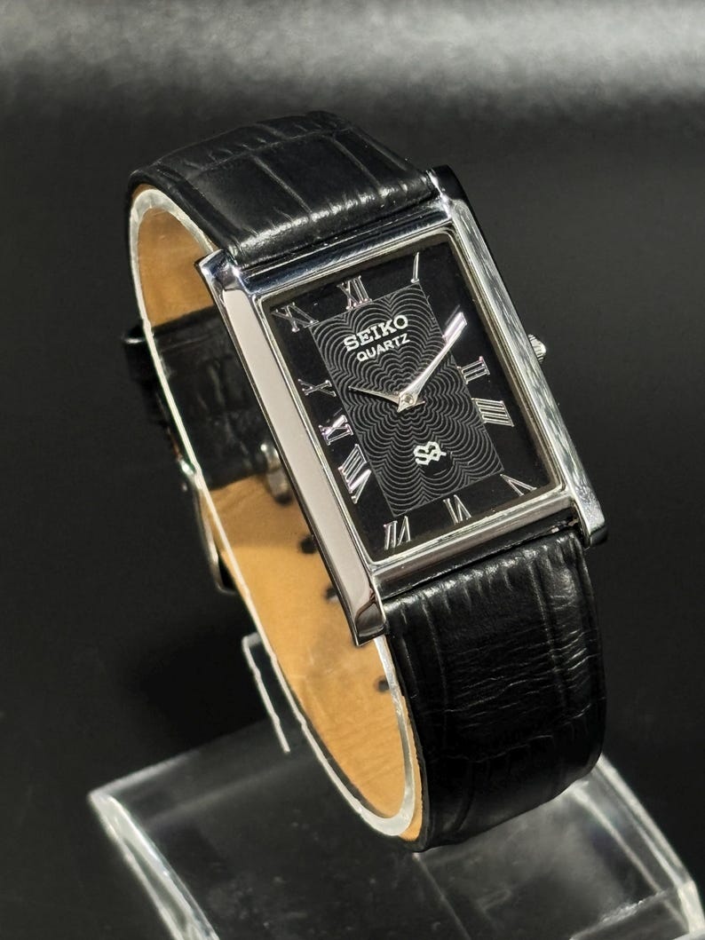 Puede incluir: Reloj Seiko Quartz rectangular con esfera negra y caja plateada. El reloj presenta n&uacute;meros romanos y una correa de cuero negro. La esfera del reloj tiene un dise&ntilde;o texturizado y el logotipo de Seiko Quartz.