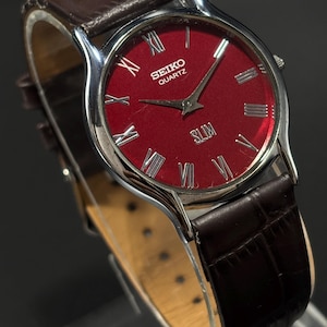 May include: A Seiko Quartz watch with a red dial and silver-tone case. The watch features Roman numeral hour markers and a brown leather strap. The word "SLIM" is printed on the dial.