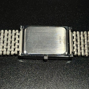 Może przedstawiać: Srebrny zegarek Seiko Quartz z prostokątną tarczą i bransoletą z siatki. Widoczny jest tył zegarka z logo i napisem Seiko. Zegarek jest umieszczony na ciemnym, teksturowanym tle.