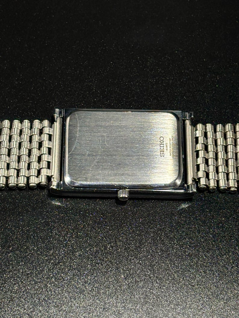 Może przedstawiać: Srebrny zegarek Seiko Quartz z prostokątną tarczą i bransoletą z siatki. Widoczny jest tył zegarka z logo i napisem Seiko. Zegarek jest umieszczony na ciemnym, teksturowanym tle.