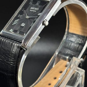 Puede incluir: Reloj Seiko Quartz rectangular con esfera negra y caja plateada. El reloj presenta n&uacute;meros romanos y un dise&ntilde;o central texturizado. Tiene una correa de cuero negro y se exhibe sobre un soporte transparente.