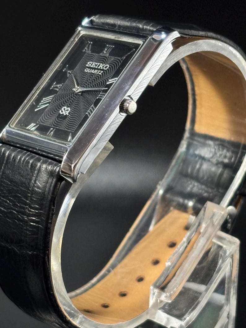 Puede incluir: Reloj Seiko Quartz rectangular con esfera negra y caja plateada. El reloj presenta n&uacute;meros romanos y un dise&ntilde;o central texturizado. Tiene una correa de cuero negro y se exhibe sobre un soporte transparente.