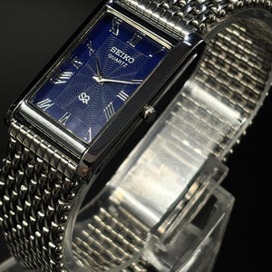 Może przedstawiać: Prostokątny zegarek Seiko Quartz z niebieską tarczą i srebrnymi rzymskimi cyframi. Zegarek ma srebrną bransoletę z siatki i srebrną kopertę. Zegarek jest prezentowany na przezroczystym stojaku.