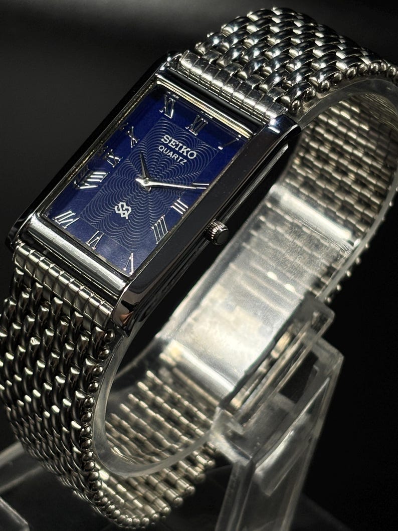 Może przedstawiać: Prostokątny zegarek Seiko Quartz z niebieską tarczą i srebrnymi rzymskimi cyframi. Zegarek ma srebrną bransoletę z siatki i srebrną kopertę. Zegarek jest prezentowany na przezroczystym stojaku.