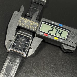 Puede incluir: Un reloj Seiko Quartz negro con una esfera rectangular y una correa de cuero negro se mide con un calibrador digital. La esfera del reloj tiene n&uacute;meros romanos y un patr&oacute;n texturizado. El calibrador muestra una medida de 27,4 mm.