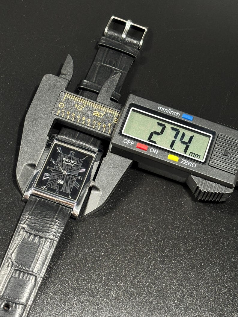 Puede incluir: Un reloj Seiko Quartz negro con una esfera rectangular y una correa de cuero negro se mide con un calibrador digital. La esfera del reloj tiene n&uacute;meros romanos y un patr&oacute;n texturizado. El calibrador muestra una medida de 27,4 mm.
