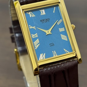 Puede incluir: Un reloj Seiko Quartz dorado con una esfera azul rectangular y números romanos dorados. El reloj tiene agujas doradas y una correa de cuero marrón. La esfera del reloj tiene el logotipo de Seiko Quartz.