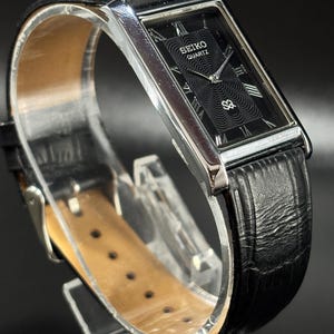 Puede incluir: Un reloj Seiko Quartz rectangular con esfera negra y agujas e indicadores plateados. El reloj tiene una correa de cuero negro y una caja plateada. La esfera muestra el logotipo de Seiko Quartz.