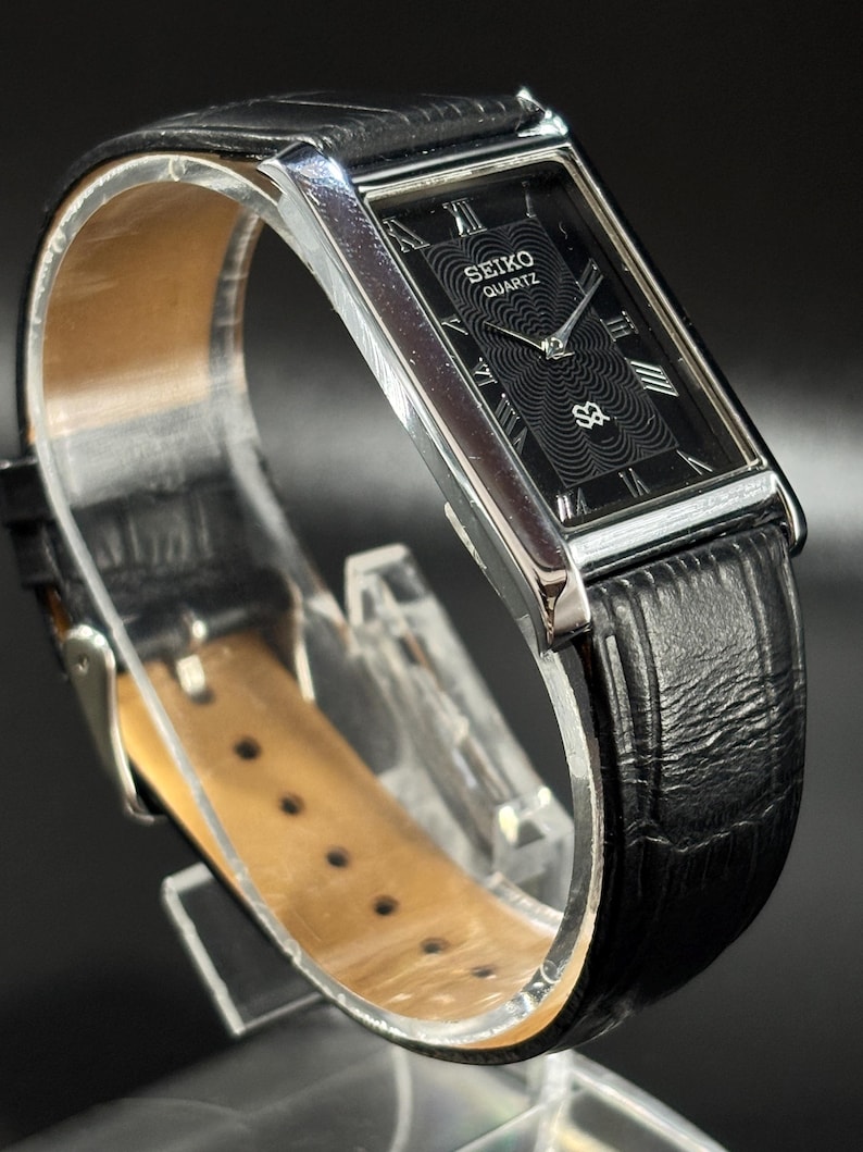 Puede incluir: Un reloj Seiko Quartz rectangular con esfera negra y agujas e indicadores plateados. El reloj tiene una correa de cuero negro y una caja plateada. La esfera muestra el logotipo de Seiko Quartz.