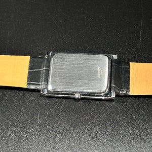 Puede incluir: Un reloj rectangular plateado con una correa de cuero negro. La esfera del reloj no es visible. La parte trasera del reloj tiene la marca "SEIKO" impresa. La correa es negra con un patr&oacute;n de cocodrilo.