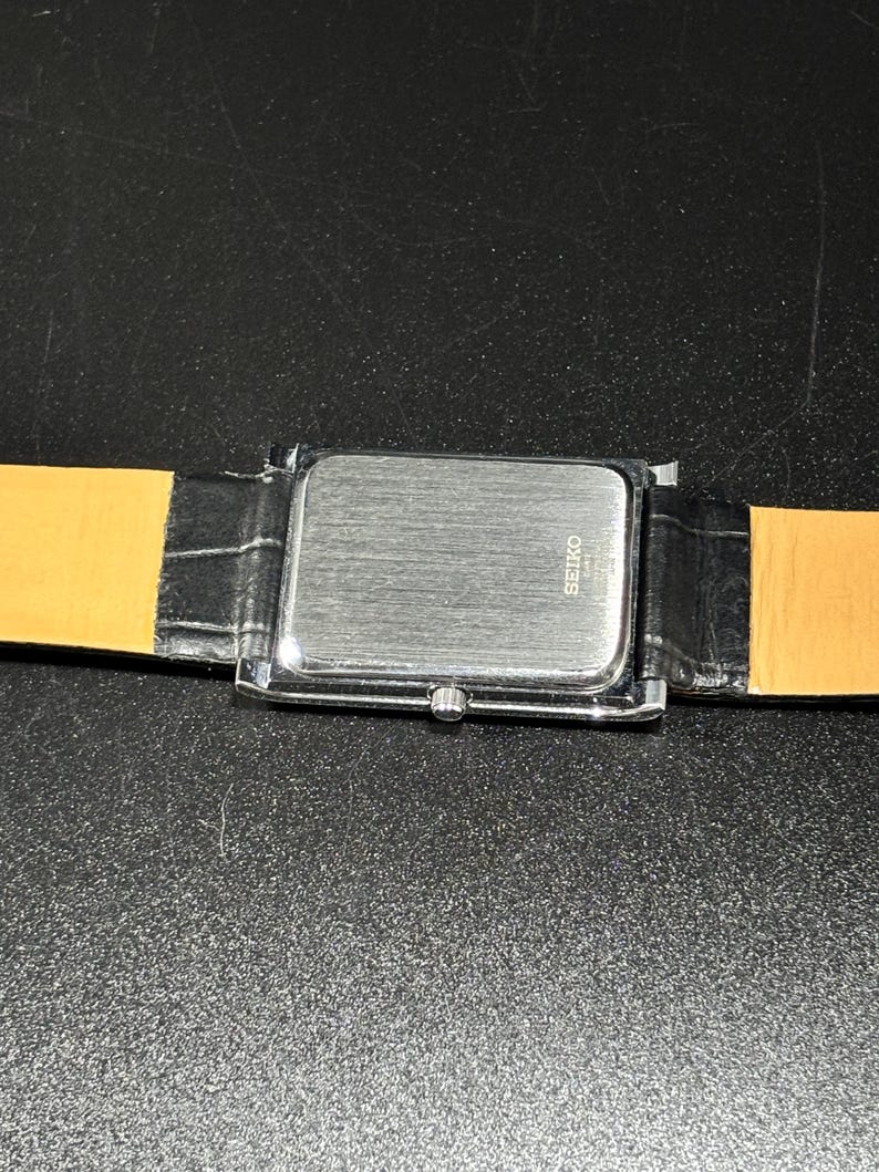 Puede incluir: Un reloj rectangular plateado con una correa de cuero negro. La esfera del reloj no es visible. La parte trasera del reloj tiene la marca "SEIKO" impresa. La correa es negra con un patr&oacute;n de cocodrilo.