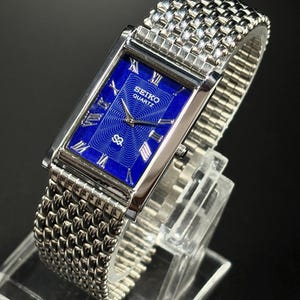 Może przedstawiać: Prostokątny zegarek Seiko Quartz z żywą niebieską tarczą i srebrnymi rzymskimi cyframi. Zegarek posiada srebrną bransoletę z siatki i jest prezentowany na przezroczystym stojaku. Tarcza zegarka ma logo Seiko Quartz.