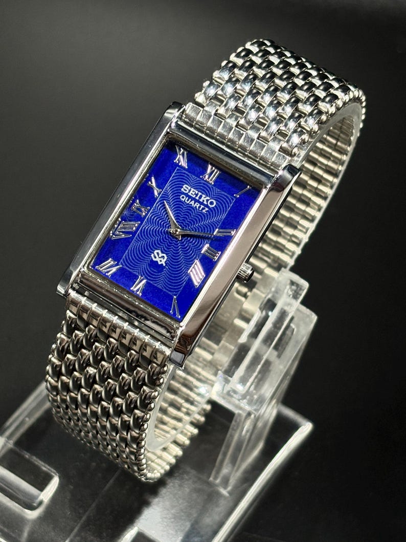 Może przedstawiać: Prostokątny zegarek Seiko Quartz z żywą niebieską tarczą i srebrnymi rzymskimi cyframi. Zegarek posiada srebrną bransoletę z siatki i jest prezentowany na przezroczystym stojaku. Tarcza zegarka ma logo Seiko Quartz.