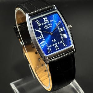 May include: A rectangular Seiko Quartz watch with a blue dial and silver hands and markers. The watch has a black leather strap and a silver case. Roman numerals mark the hours. The watch is displayed on a clear stand.
