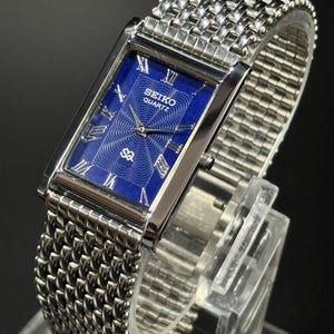 Może przedstawiać: Prostokątny zegarek Seiko Quartz z niebieską tarczą i srebrnym metalowym paskiem. Tarcza zegarka posiada rzymskie cyfry i wz&oacute;r. Zegarek jest prezentowany na przezroczystym stojaku na ciemnym tle.