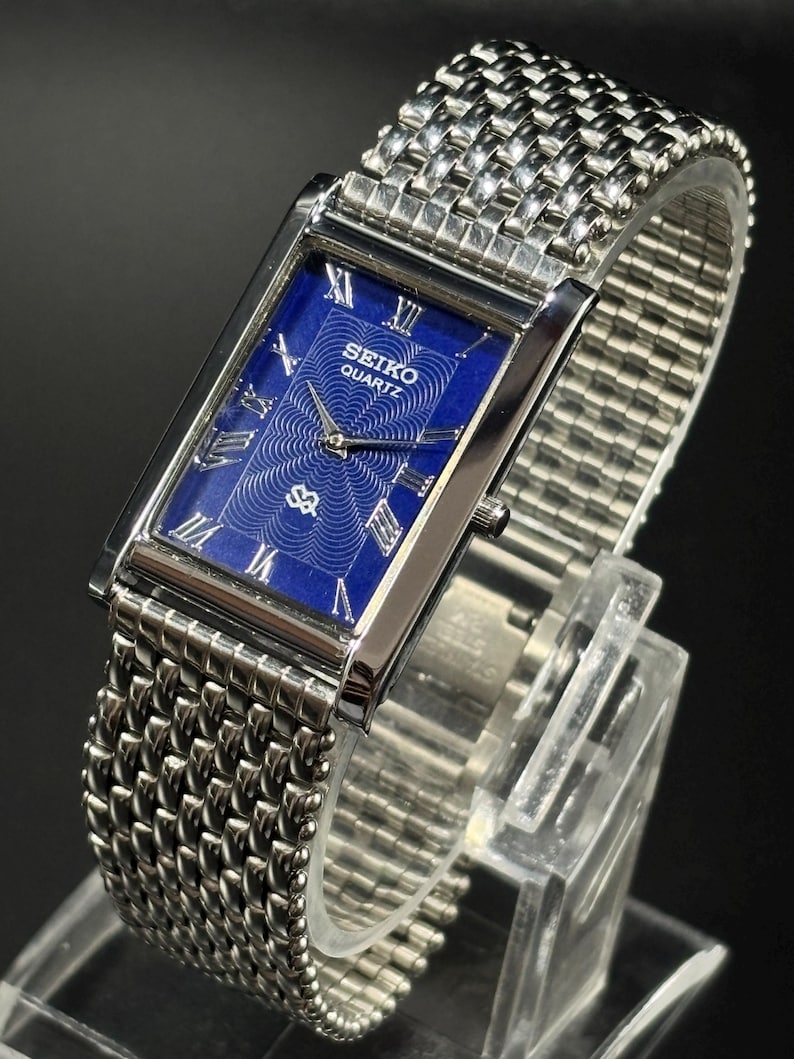 Może przedstawiać: Prostokątny zegarek Seiko Quartz z niebieską tarczą i srebrnym metalowym paskiem. Tarcza zegarka posiada rzymskie cyfry i wz&oacute;r. Zegarek jest prezentowany na przezroczystym stojaku na ciemnym tle.