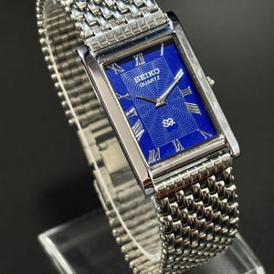 Puede incluir: Reloj Seiko Quartz rectangular con correa de metal plateado. La esfera azul vibrante muestra números romanos y el logotipo de Seiko Quartz. La correa tiene un diseño tejido.