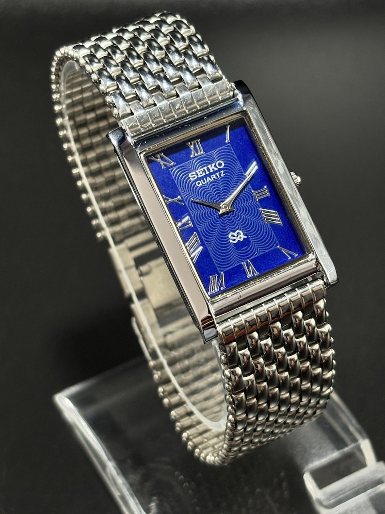 Może przedstawiać: Prostokątny zegarek Seiko Quartz ze srebrną metalową bransoletą. Tarcza zegarka jest w żywym niebieskim kolorze z rzymskimi cyframi i logo Seiko Quartz. Bransoleta ma pleciony wz&oacute;r.