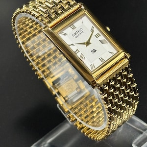 May include: A gold-coloured Seiko Quartz watch with a rectangular face and a woven metal strap. The watch face is white with black Roman numerals and hands. The watch is displayed on a clear stand against a dark background.
