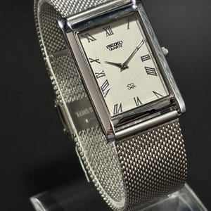Puede incluir: Un reloj Seiko Quartz plateado con esfera rectangular y correa de malla. La esfera es blanca con números romanos y agujas negras. La correa es de acero inoxidable tejido. El reloj se exhibe sobre un soporte transparente.