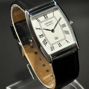 May include: A rectangular Seiko Quartz watch with a white face, black Roman numerals, and a black leather band. The watch has a silver-colored bezel and a small seconds dial. The watch is displayed on a clear stand.