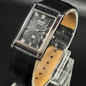 Puede incluir: Un reloj Seiko Quartz rectangular con esfera negra y caja plateada. El reloj presenta marcadores de hora con n&uacute;meros romanos y un dise&ntilde;o central texturizado. Tiene una correa de cuero negro.