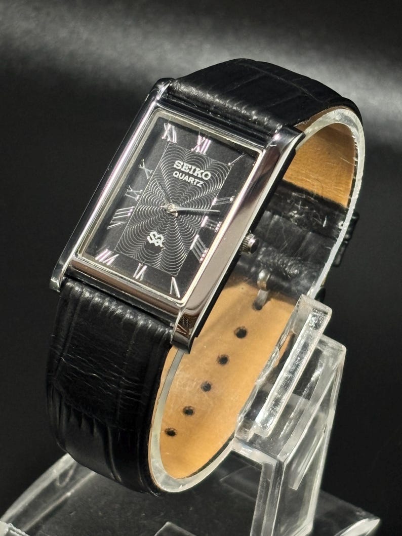 Puede incluir: Un reloj Seiko Quartz rectangular con esfera negra y caja plateada. El reloj presenta marcadores de hora con n&uacute;meros romanos y un dise&ntilde;o central texturizado. Tiene una correa de cuero negro.