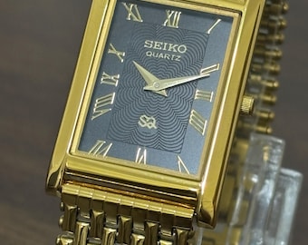 May include: A gold-toned Seiko Quartz wristwatch with a rectangular face and a matching linked band. The watch face is dark gray with gold Roman numerals and hands. The watch is displayed on a clear stand.