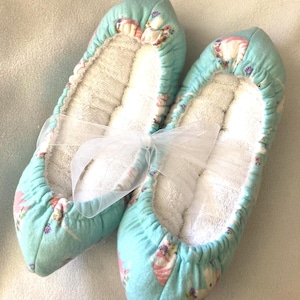 May include: A pair of light blue slippers featuring a repeating unicorn and floral pattern. The slippers have a gathered elastic opening and a white, textured interior lining. A sheer white ribbon is tied around the center of the pair.