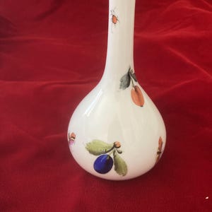 May include: A white ceramic vase with a gold rim and a floral design featuring plums, leaves, and berries. The vase has a narrow neck and a rounded body.