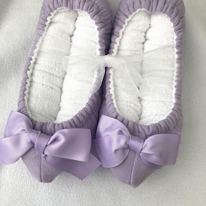 Op de afbeelding: Een paar lavendelkleurige pantoffels met witte badstof binnenkant en bijpassende lavendelkleurige strikken. De pantoffels zijn samengebonden met een doorschijnend wit lint. Ontworpen voor gebruik binnenshuis.