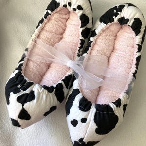 May include: A pair of slip-on slippers featuring a black and white cow print pattern on the exterior and a soft, light pink textured fabric lining. The two slippers are tied together with a sheer white ribbon bow across the center.