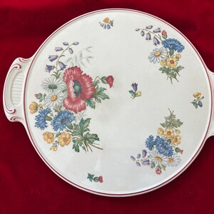 Op de afbeelding: Een witte keramische serveerschaal met twee handvatten. De schaal is versierd met een bloemenpatroon met rode klaprozen, blauwe korenbloemen, gele madeliefjes en groene bladeren.