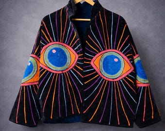 Könnte beinhalten: Schwarze Samtjacke mit gestickten Augendesigns in leuchtenden Farben. Die Jacke hat große, stilisierte Augen auf der Vorderseite und den Ärmeln, mit strahlenförmigen Linien in Orange, Rosa, Lila und Blau. Die Jacke hängt an einem Holzkleiderbügel.