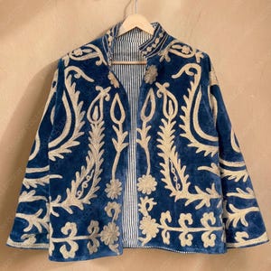 May include: A blue velvet jacket with gold embroidery. The jacket has a mandarin collar and a striped lining. The gold embroidery features floral and leaf patterns. The jacket is hanging on a wooden hanger.