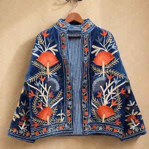 Puede incluir: Una chaqueta de terciopelo azul profundo con cuello alto y frente abierto. La prenda está decorada con intrincados bordados florales en hilos de color naranja vibrante, blanco y gris. El forro interior muestra un patrón de rayas verticales azul y blanco.