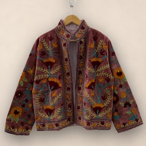 May include: A dusky pink velvet jacket with detailed floral embroidery in yellow, orange, and teal. The jacket has a high collar and a striped lining. The embroidery is focused around the collar, front, and sleeves.