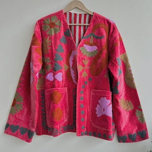 May include: A bright pink velvet jacket adorned with detailed floral and leaf embroidery in green, brown, and purple. The jacket has two front pockets and a striped lining, displayed on a wooden hanger.