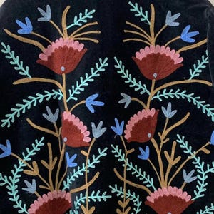 May include: Black garment with embroidered floral design. The embroidery features maroon, pink, blue, and gold-colored flowers and leaves. The garment has a velvet-like texture and a detailed border along the bottom.
