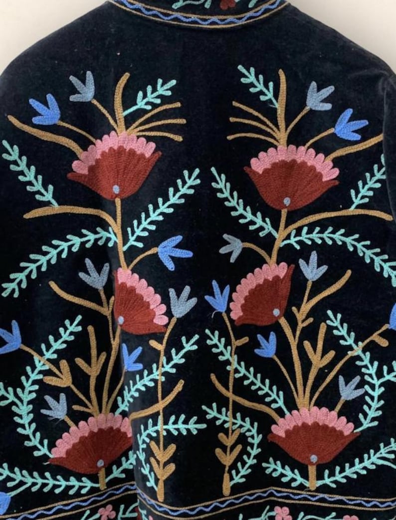 May include: Black garment with embroidered floral design. The embroidery features maroon, pink, blue, and gold-colored flowers and leaves. The garment has a velvet-like texture and a detailed border along the bottom.