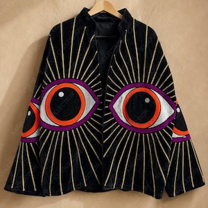 May include: Black velvet jacket featuring gold vertical stripes and large embroidered eye designs in orange, purple, and white. The jacket has a high collar and is hanging on a wooden hanger.