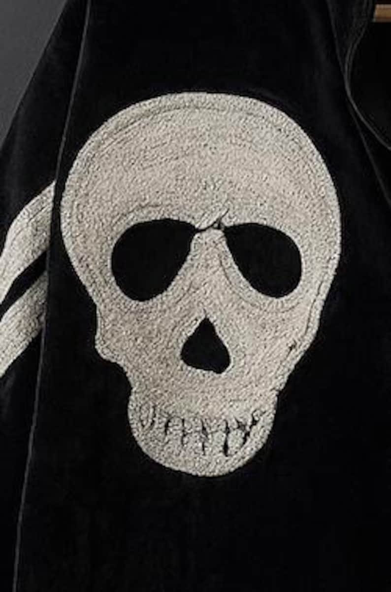 May include: Close-up of a black jacket featuring a large, textured, off-white skull emblem on the front. The skull has prominent eye sockets and a detailed jawline. A white stripe is visible on the left side of the jacket.