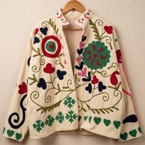 以下が含まれることがあります： 鮮やかな多色の花柄と幾何学模様の刺繍が施されたオフホワイトのジャケット。フロントと袖には赤、緑、ピンク、濃紺の糸で模様が描かれており、スタンドカラーと長袖が特徴です。