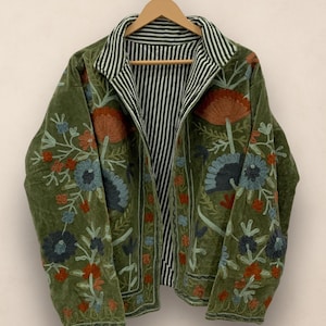 Puede incluir: Chaqueta de terciopelo verde oliva con bordados florales en tonos azules, naranjas y verdes. La chaqueta tiene forro y cuello a rayas. Cuelga de una percha de madera sobre un fondo neutro.