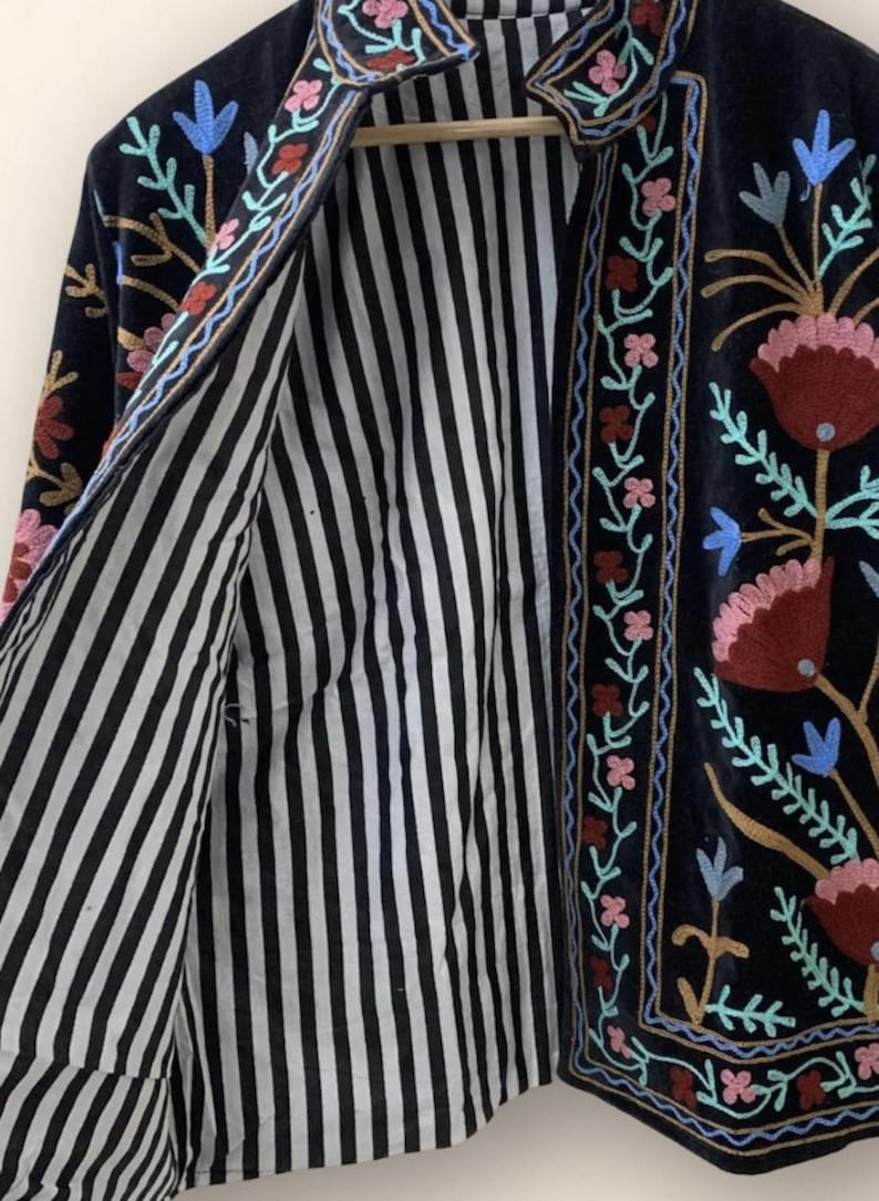 May include: A dark navy blue jacket with floral embroidery in pink, red, blue, and yellow. The jacket's interior features black and white vertical stripes. The embroidery is along the edges and collar.